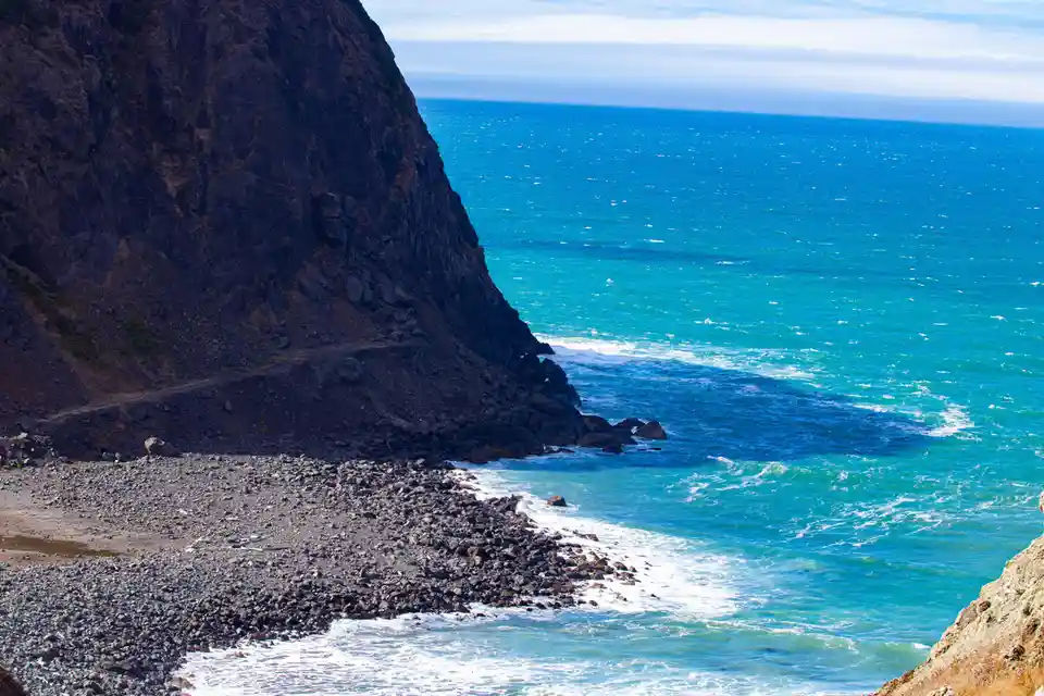 A scenic coastal view shows a steep, rocky cliffside meeting turquoise ocean waves crashing onto a pebbly shore under a…