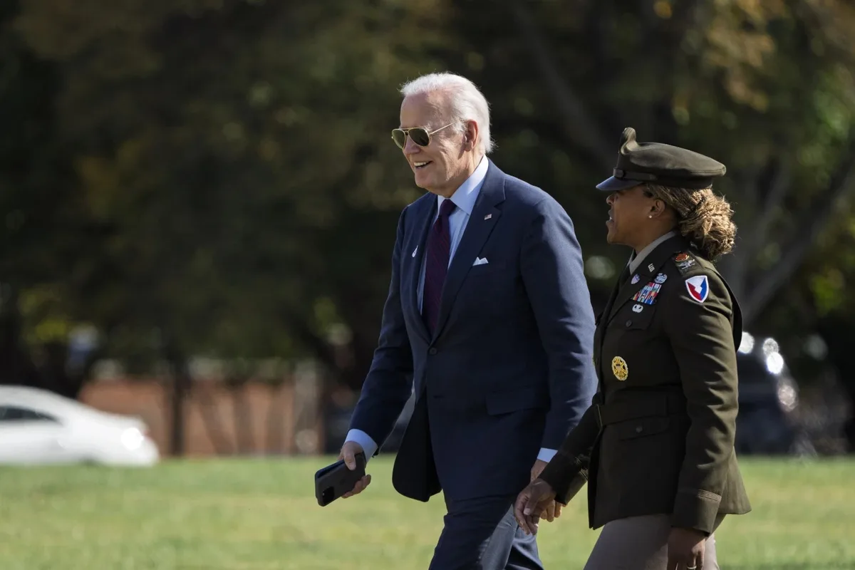 a man in a dark suit and sunglasses walks beside a woman in a military uniform, both holding hands as they stroll outdoors…