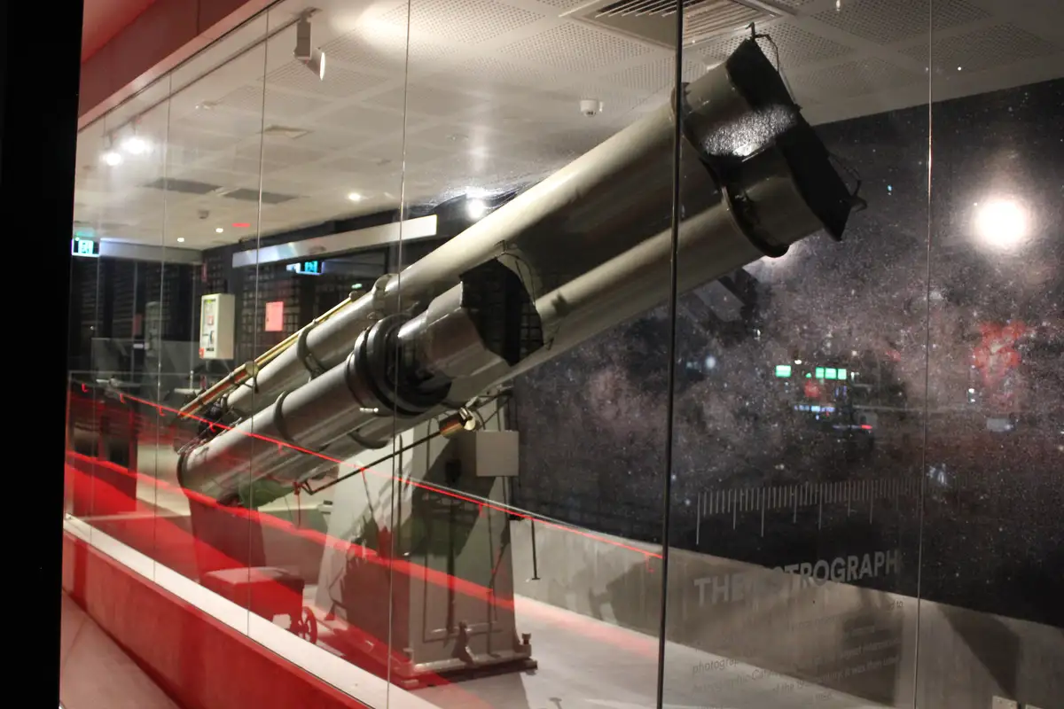 A large, silver, tube-shaped telescope called 'THE ASTROGRAPH' is displayed inside a glass case in a museum, with a starry…