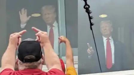 A man in a red shirt and black cap points upward with both index fingers toward a glass window. Behind the glass, Donald…