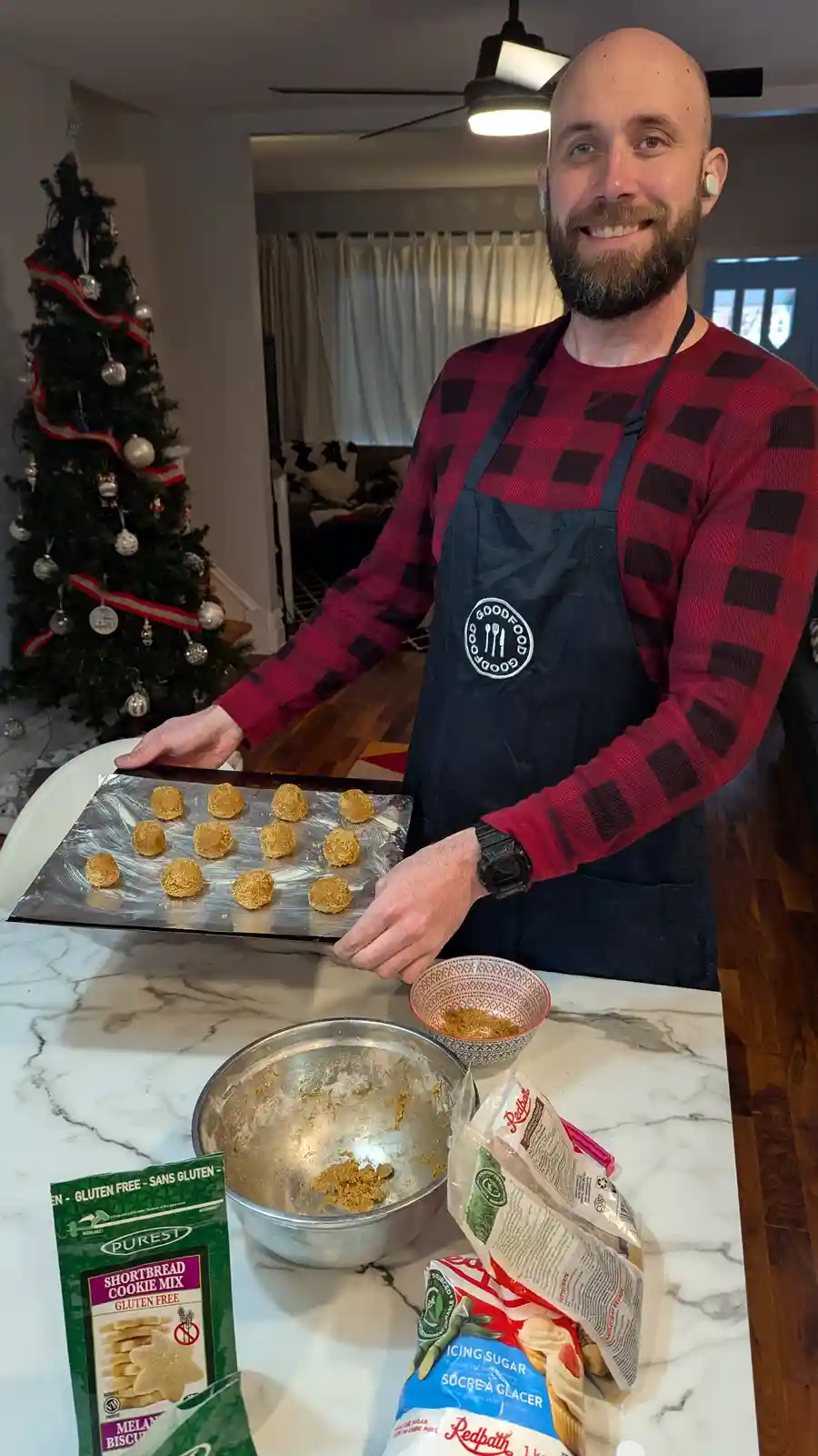 A smiling bald man with a beard, wearing a red plaid shirt and a black apron, holds a baking sheet with round, golden-brown…