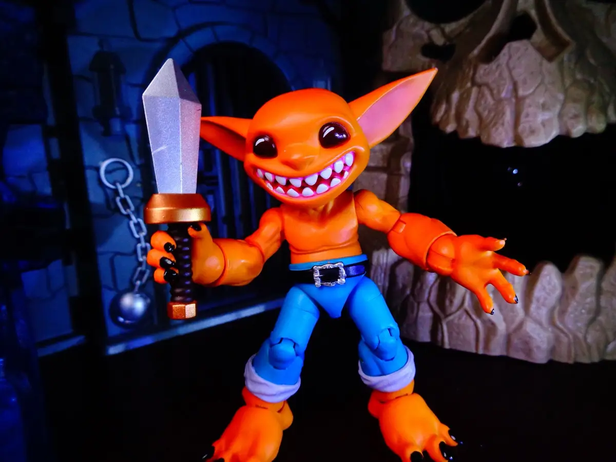 An orange, goblin-like action figure with large ears, sharp teeth, and black fingernails holds a sword while posing in front…