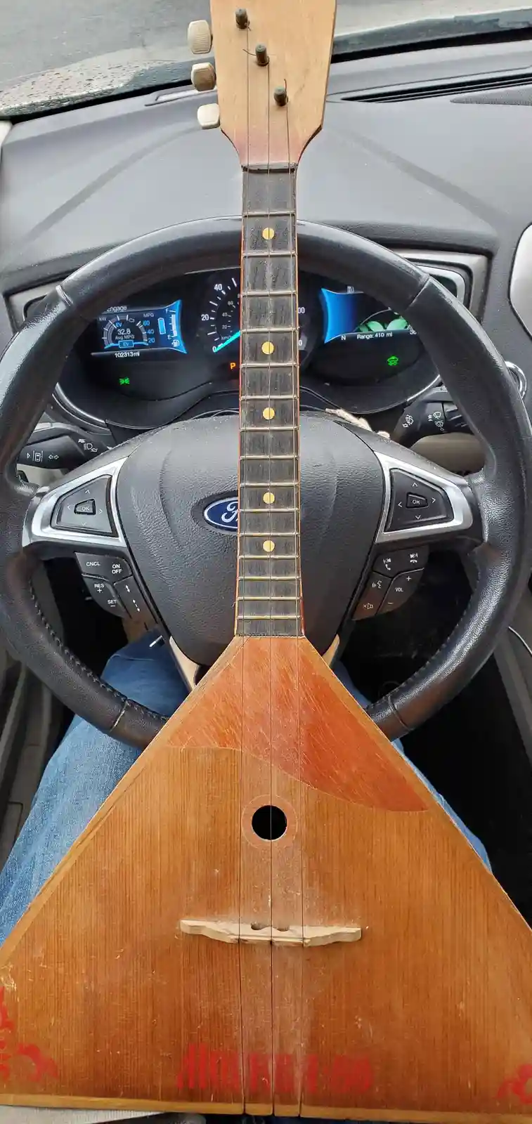A triangular balalaika is positioned vertically in the center of a car's steering wheel, with its neck extending upward…