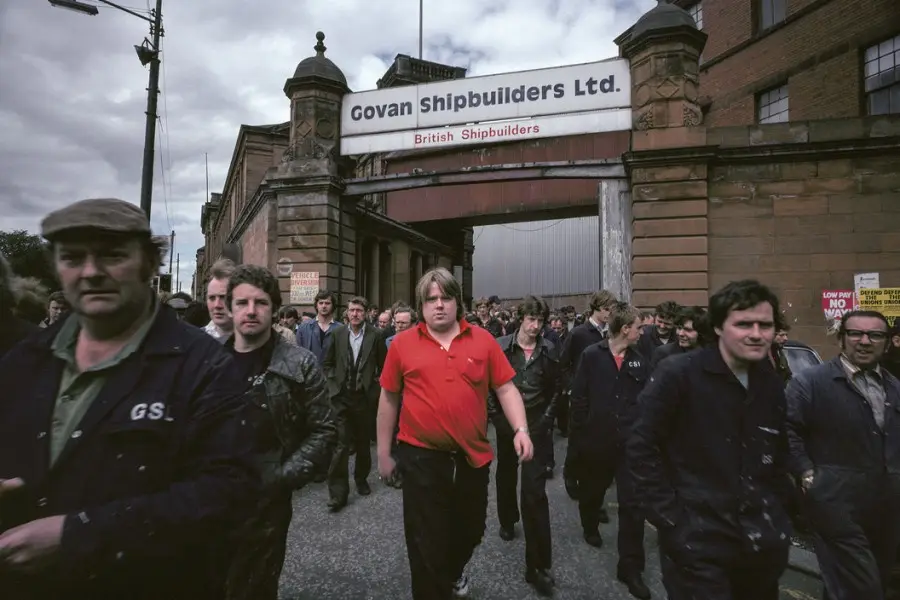 A historical photograph shows a large group of men, mostly in work clothes, walking out of the gates of Govan Shipbuilders…