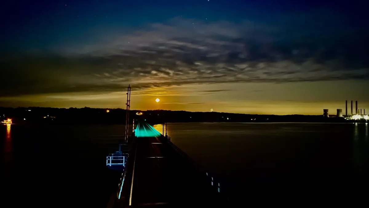 A nighttime photograph shows a long, dark pier extending over water toward a horizon where a bright moon hangs low in a…