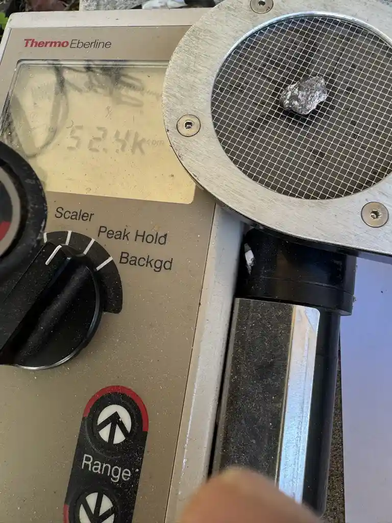 A close-up photo shows a small, rough rock resting on a metal grid inside a Thermo Eberline radiation detector. The device's…