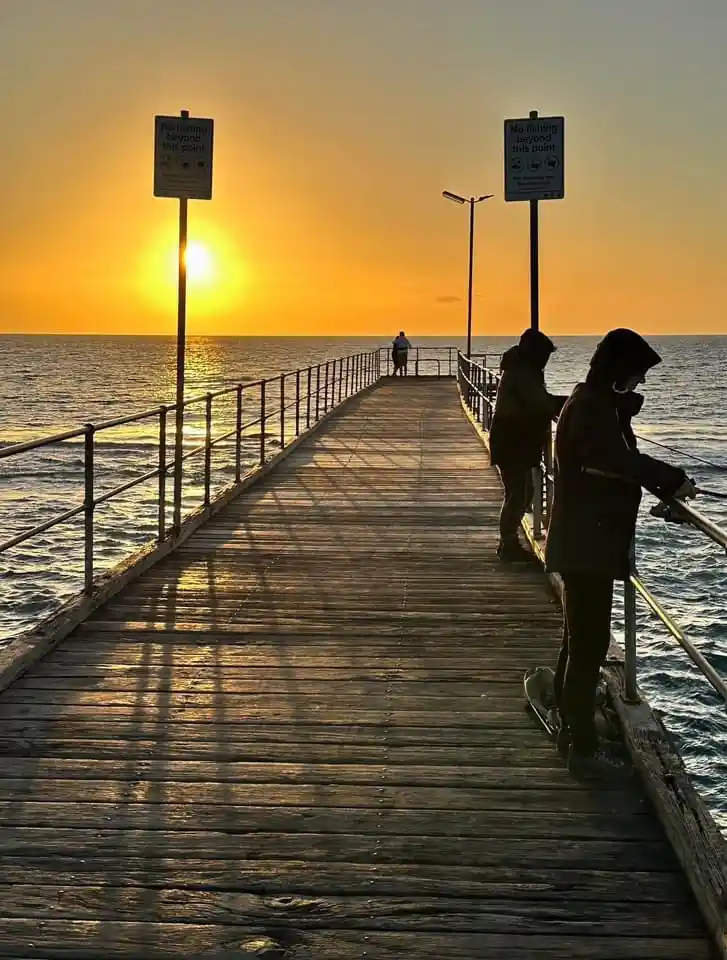 A serene sunset scene on a wooden pier with two people fishing near the end. Two identical signs on posts read 'No fishing…