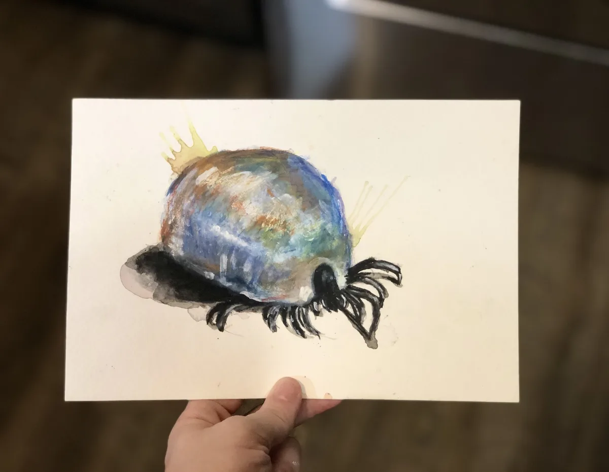A hand holds up a watercolor painting of a pearl with a small yellow crown on top and black eyelashes drawn around its lower…