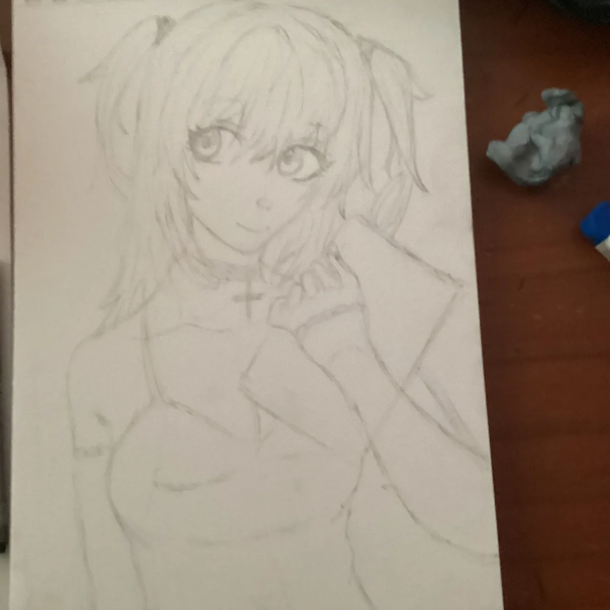 A pencil sketch of an anime-style girl with pigtails and a cross necklace, drawn on white paper. The drawing is unfinished…