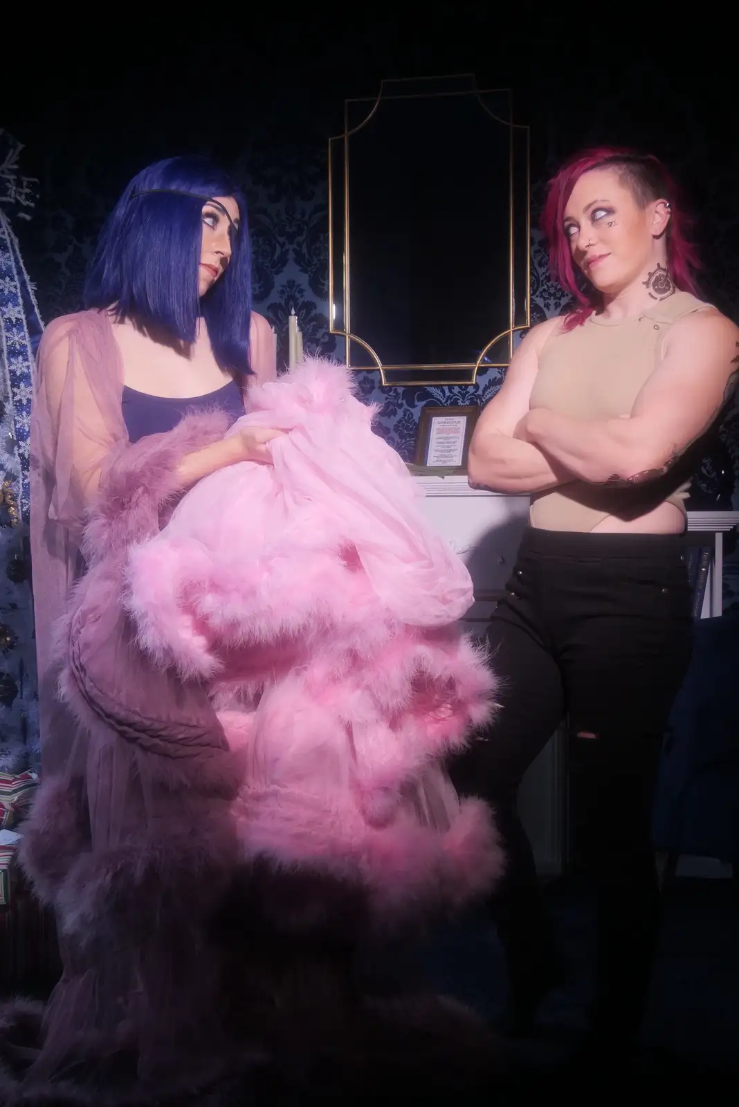 Two women stand in a dimly lit room with dark patterned wallpaper. The woman on the left, with blue hair and wearing a sheer…