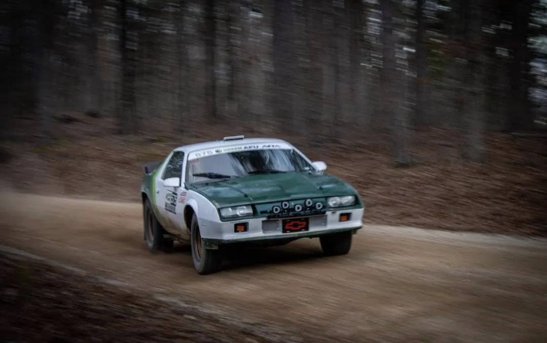 A green and white rally car speeds down a dirt road through a forest, with motion blur indicating high speed. The front…