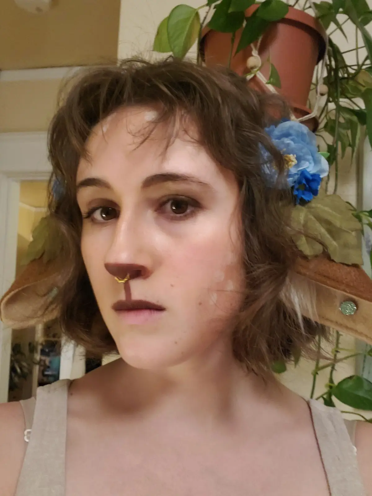 A person with short brown hair and a nose ring looks at the camera with a neutral expression. They have white face paint on…