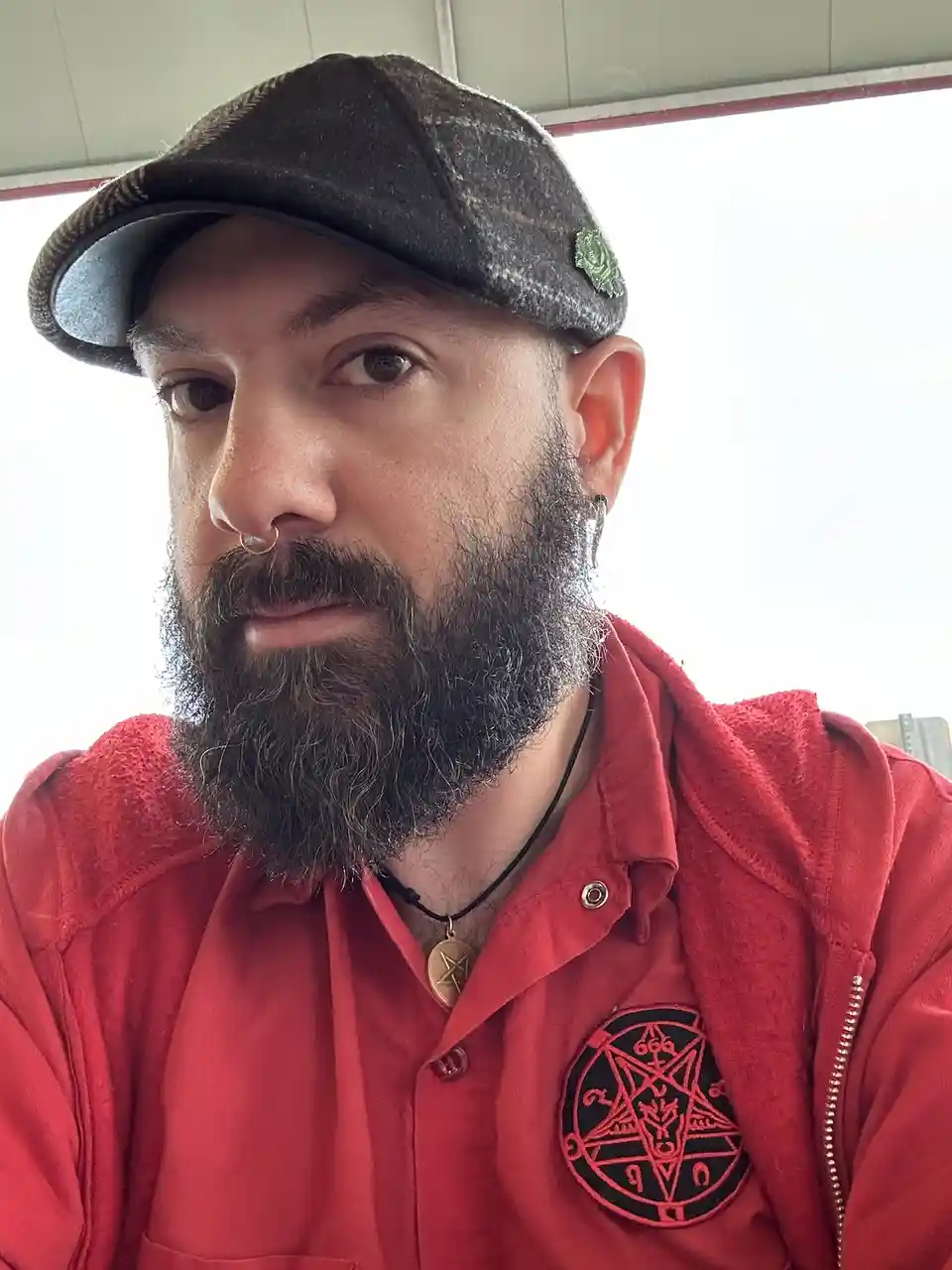 A man with a beard, nose ring, and earring wears a red jacket with a pentagram patch and a pentagram pendant. He also wears…