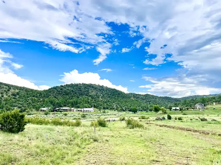A scenic rural landscape featuring rolling green hills, scattered houses, and a bright blue sky with white clouds. The…