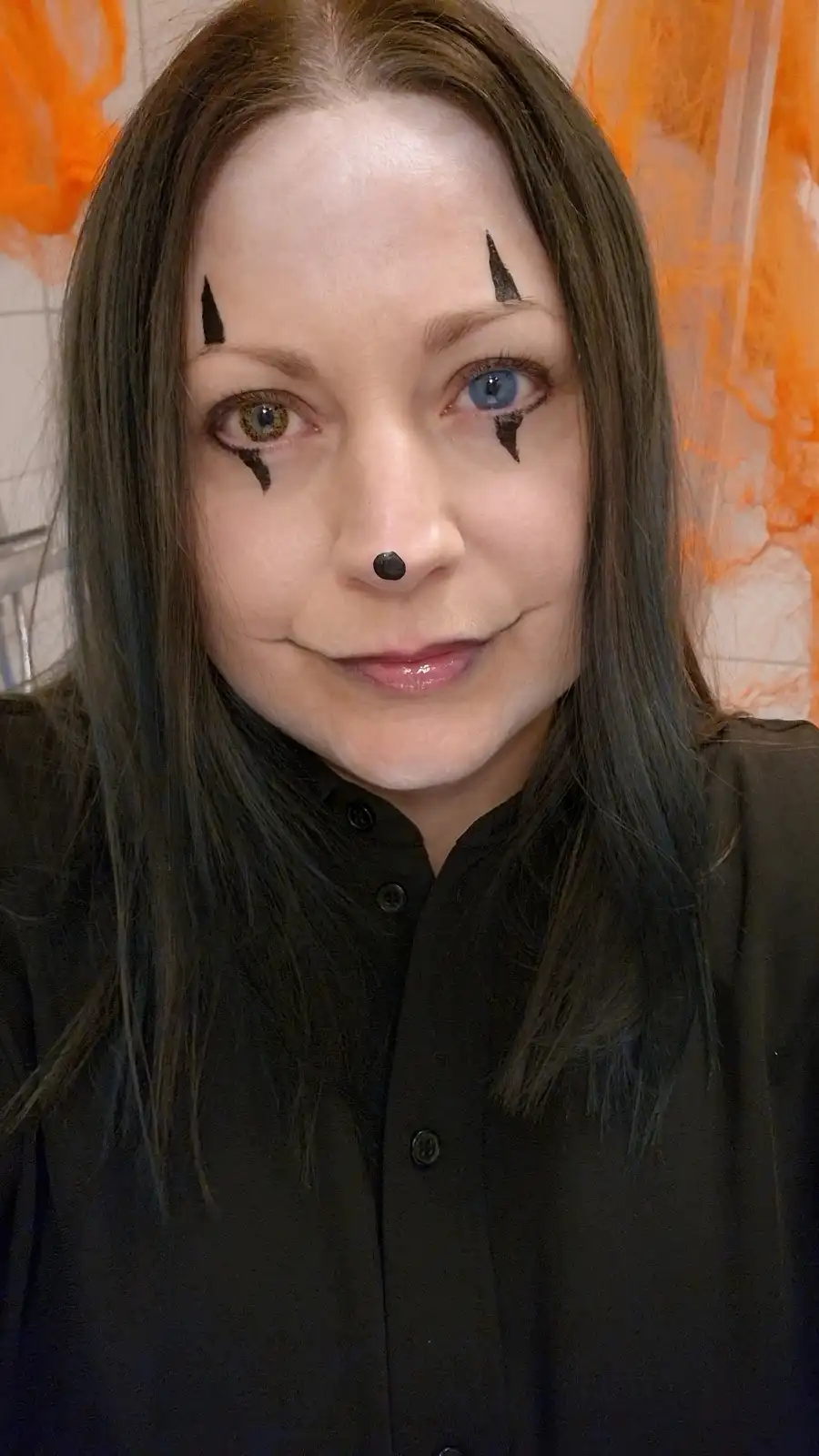 A person with dark hair and a black button-up shirt poses with Halloween clown makeup: black triangular markings above and…