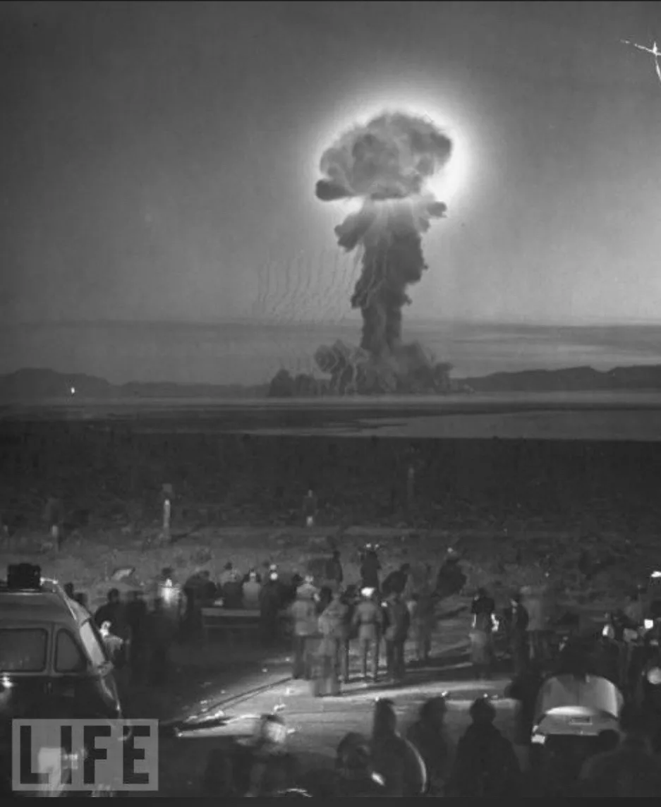 A black-and-white historical photograph shows a massive nuclear mushroom cloud rising over a desert landscape, with a crowd…