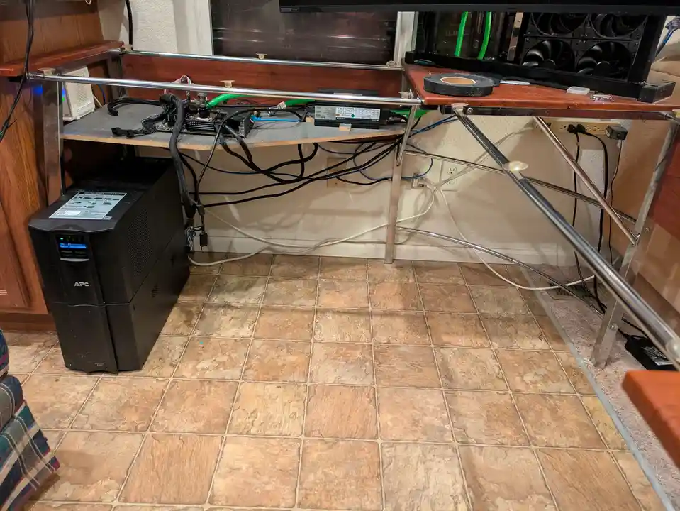A cluttered home setup shows a makeshift desk with a mining rig, power supply, and tangled cables. An APC uninterruptible…