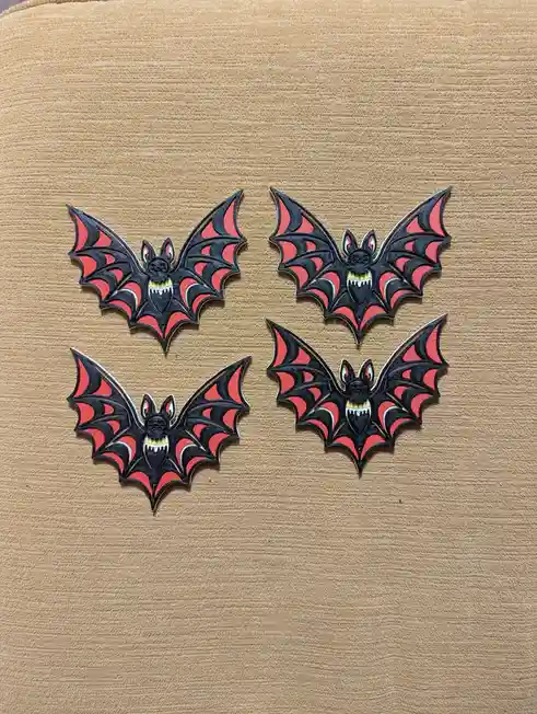 Four identical black and red bat-shaped enamel pins with sharp teeth and bat ears are arranged in a diamond pattern on a…
