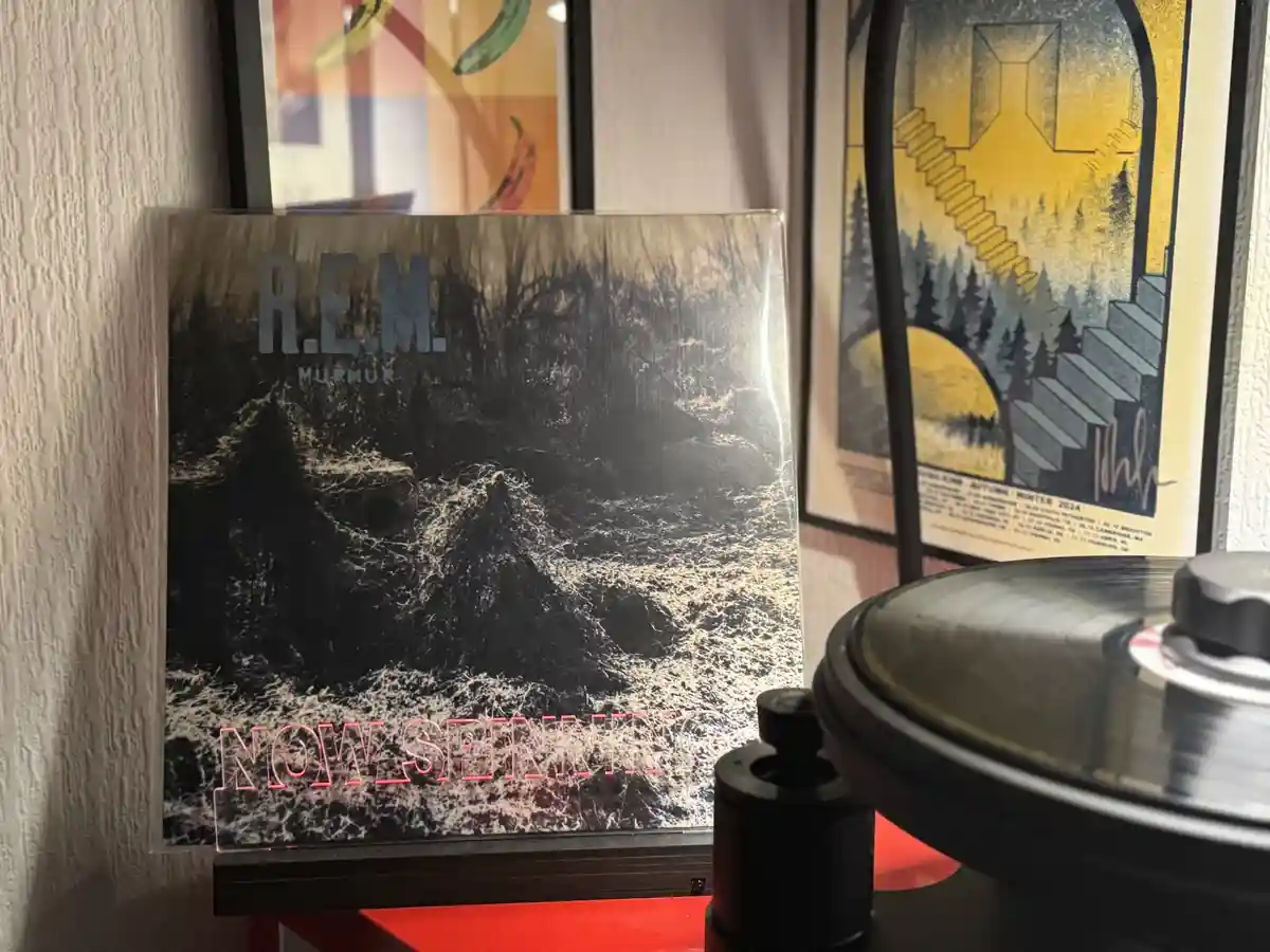 A vinyl record of R.E.M.'s album 'Murmur' is shown standing upright next to a record player with a record on it. The album…