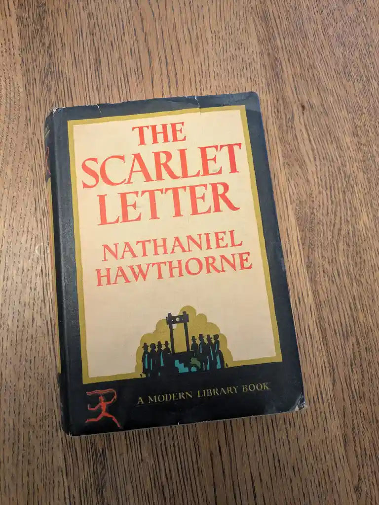 A physical copy of Nathaniel Hawthorne's 'The Scarlet Letter' sits on a wooden surface, showing its cover with red text and…