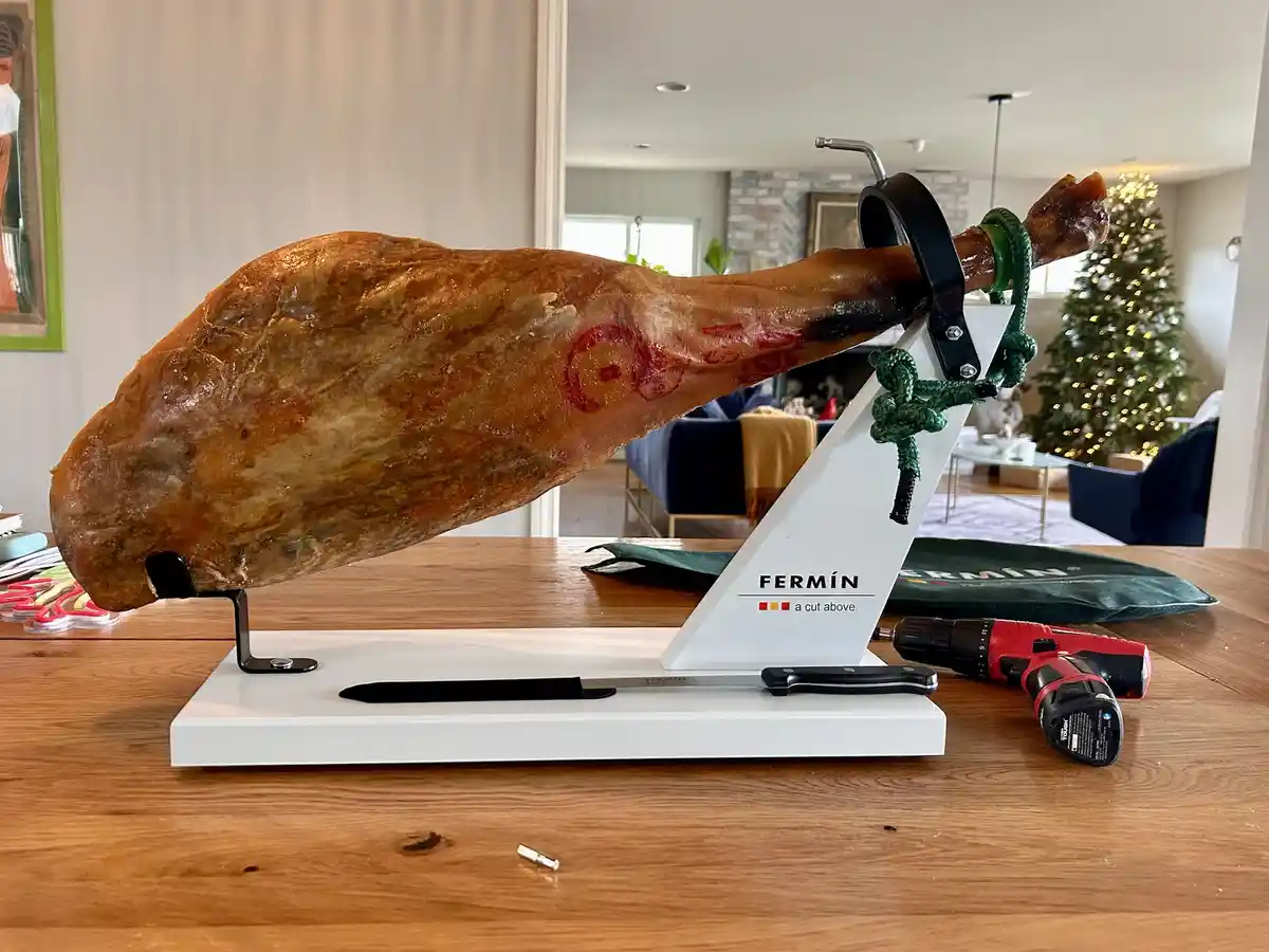 A large cured ham leg is displayed on a white 'FERMÍN a cut above' branded stand on a wooden table, with a red and black…