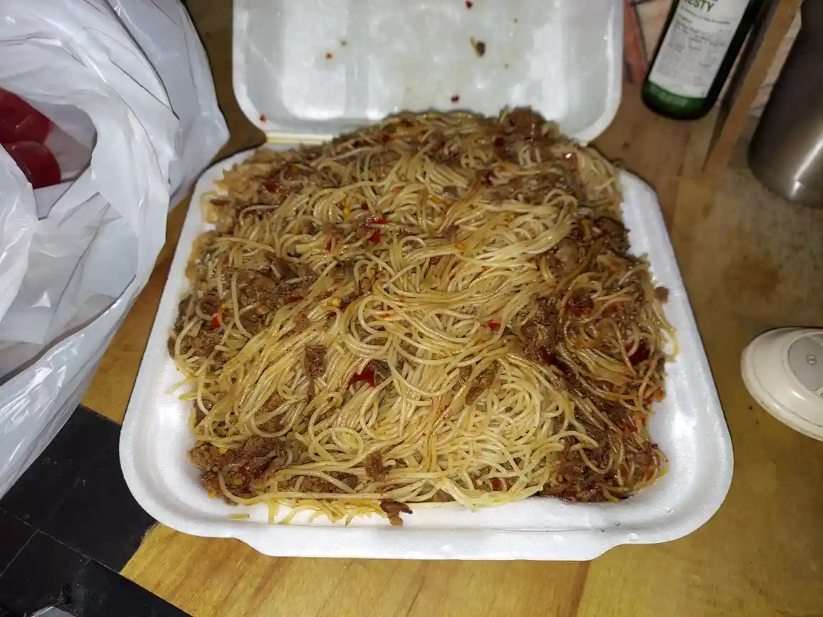 A close-up photo shows a white styrofoam takeout container filled with spaghetti mixed with ground meat and red chili…