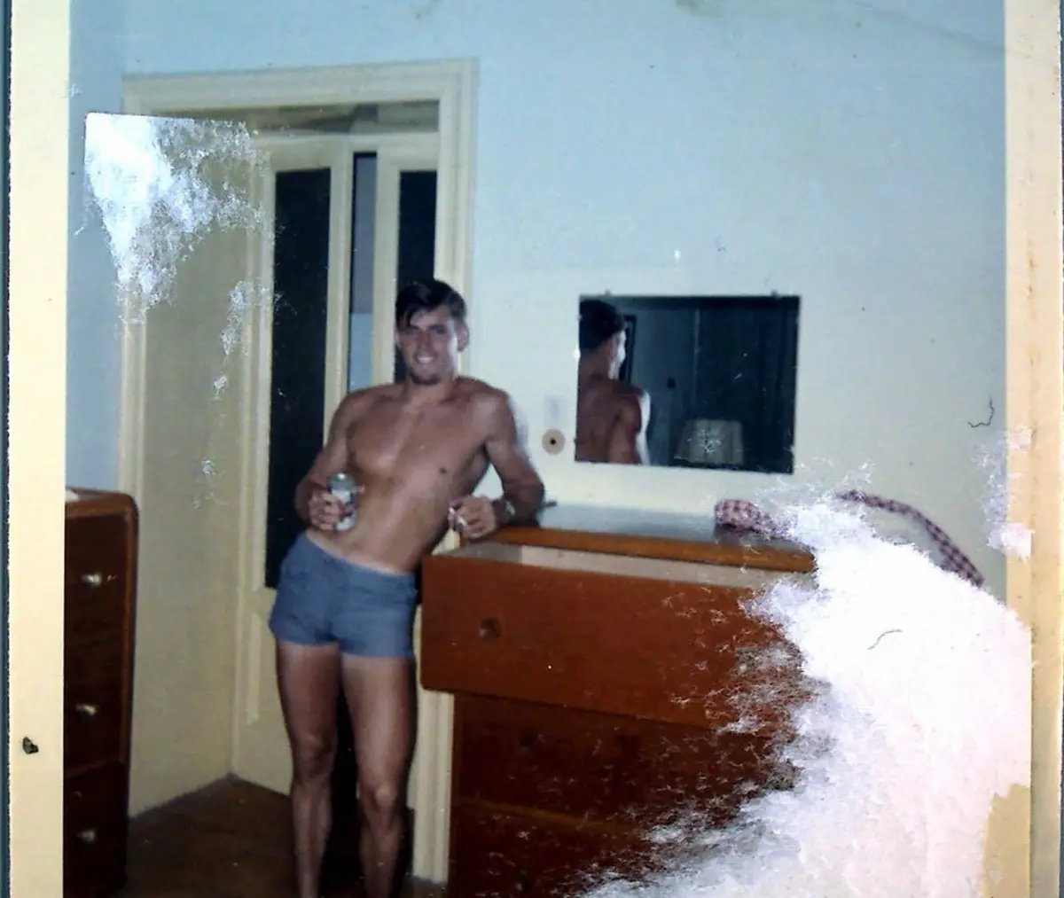 A grainy, vintage photo shows a shirtless man in blue shorts standing in a bedroom, holding a can and smiling at the camera…