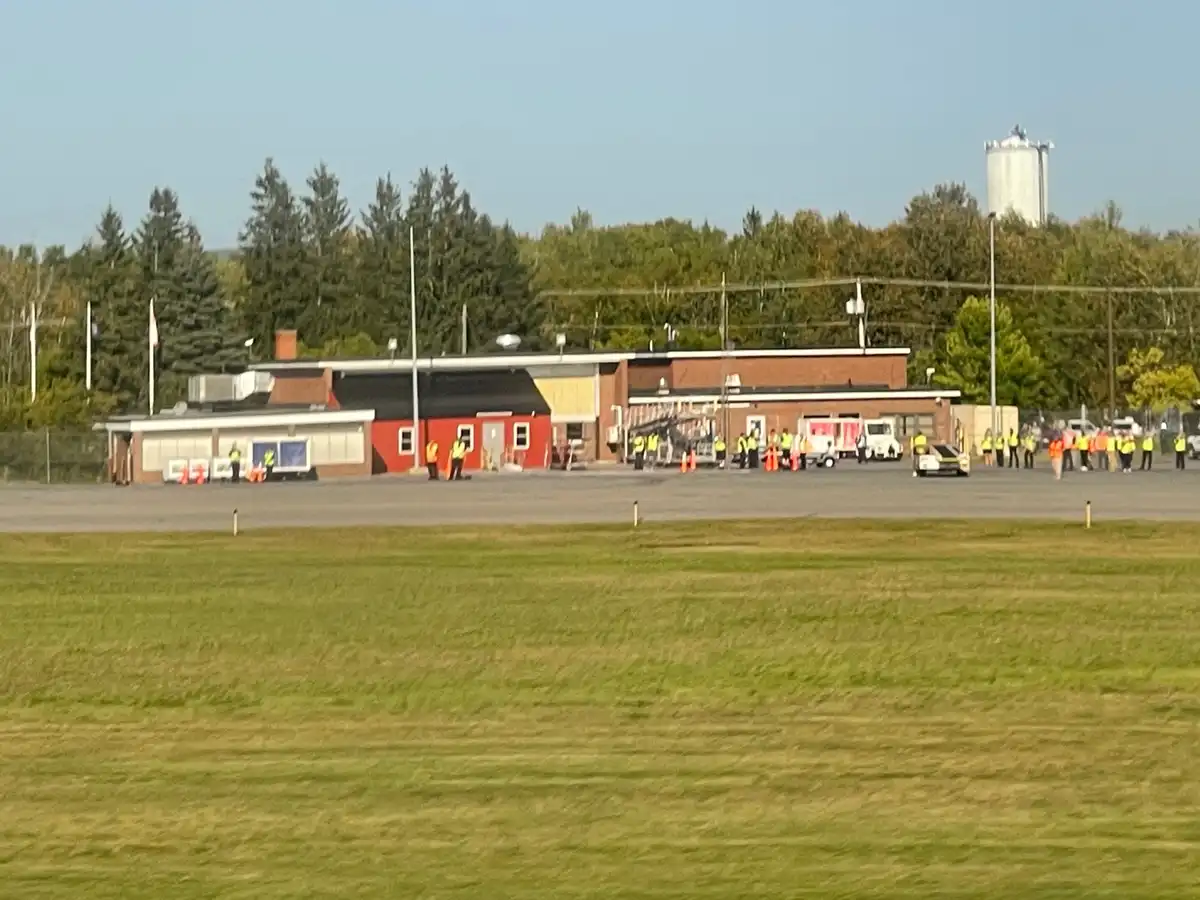 A wide shot of an airport tarmac with a red and brick building in the background, surrounded by trees and a silo. Numerous…