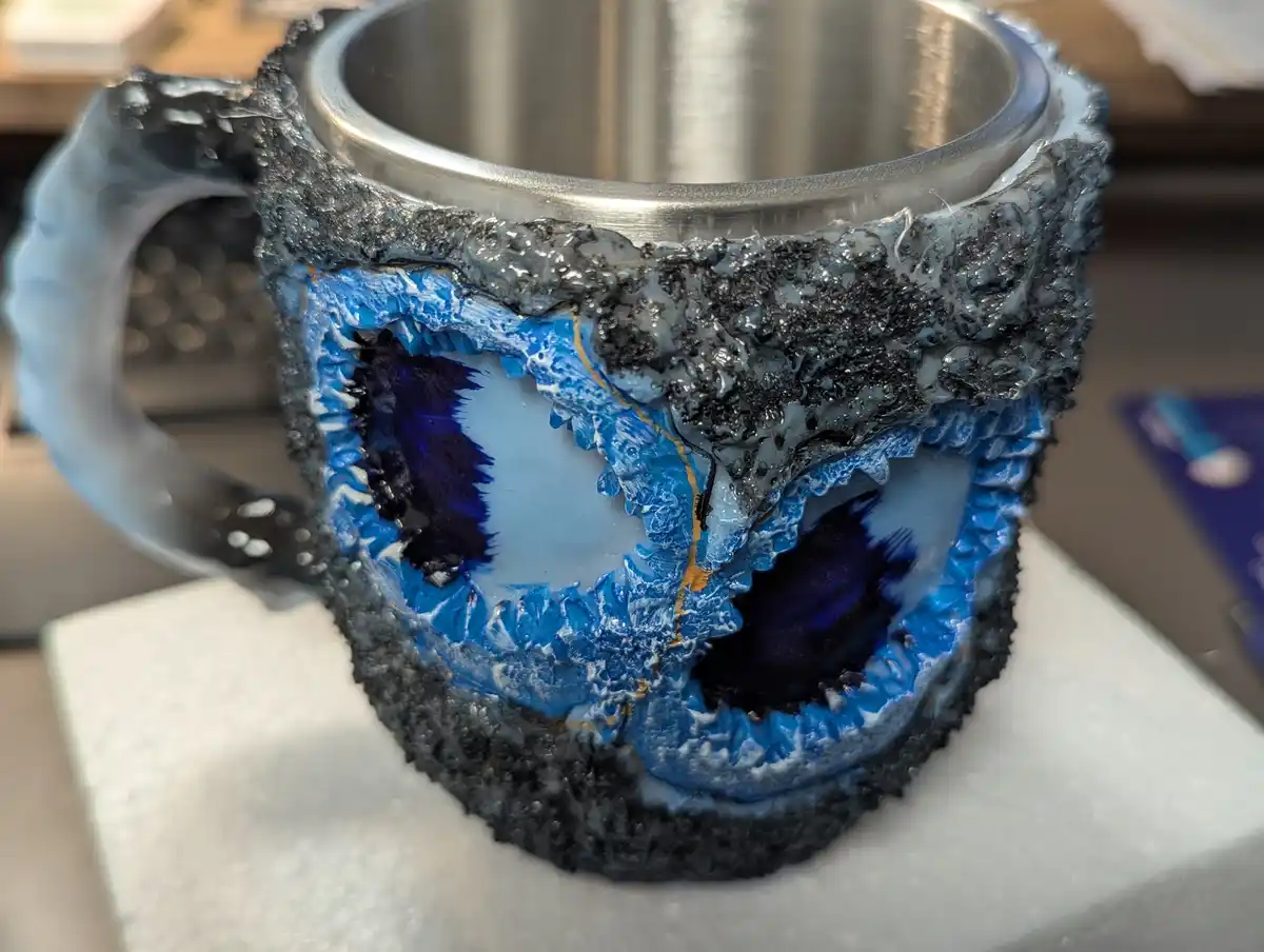 A stainless steel mug has been decorated with a blue and black geode-like resin design that looks like a cracked…