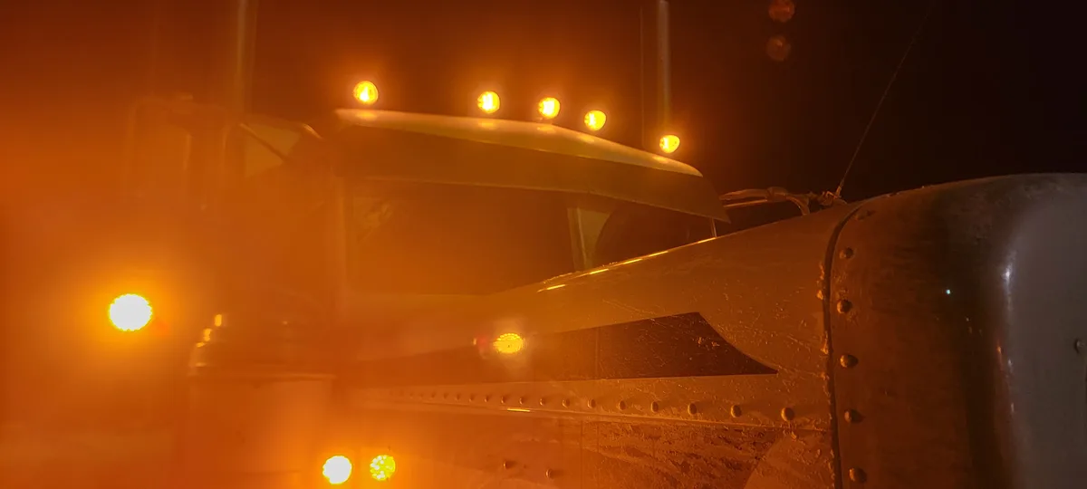 A close-up nighttime photo of a large, rugged semi-truck cab, its surface covered in frost or snow, illuminated by glowing…