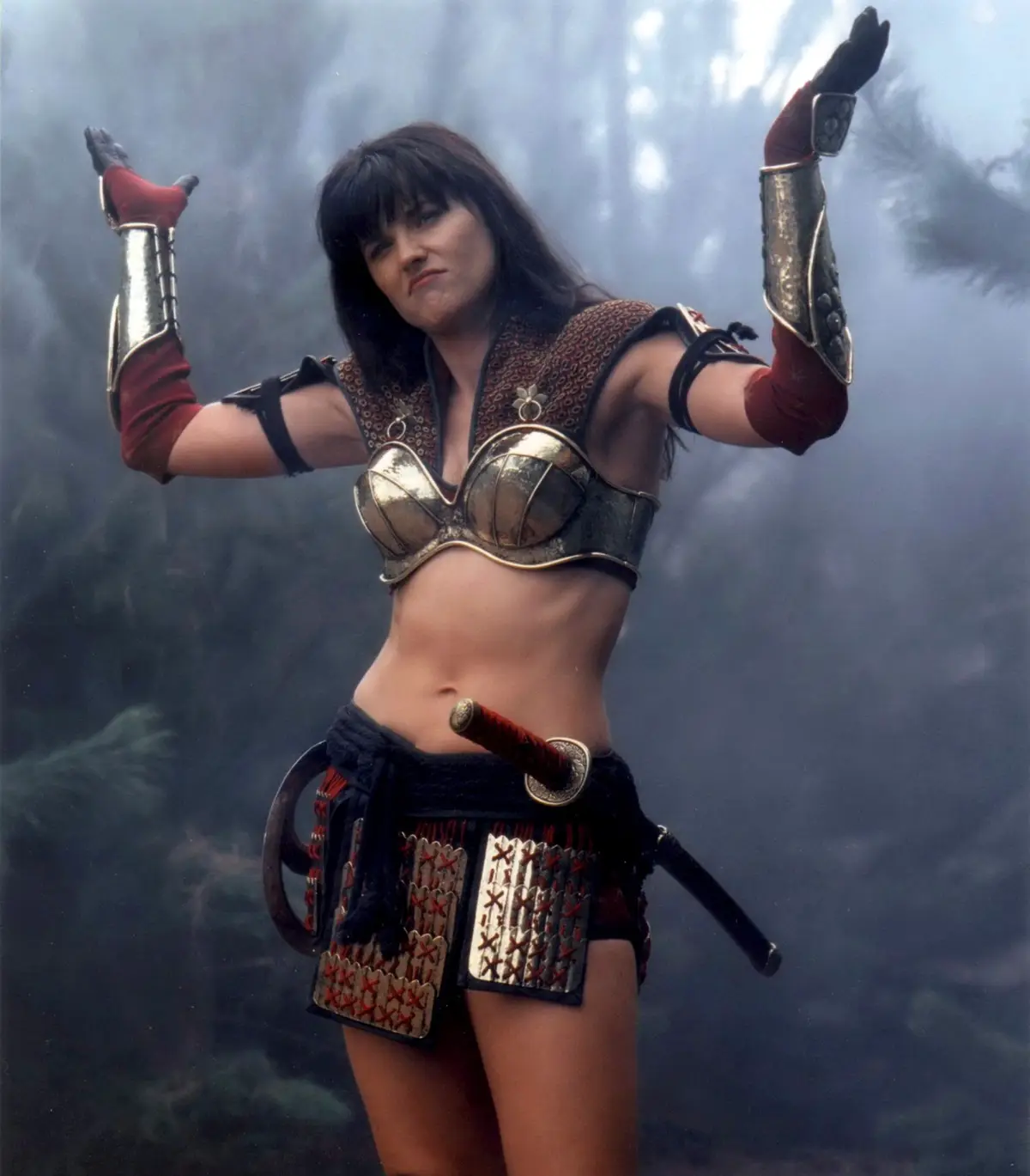 A woman in a fantasy warrior outfit with metallic arm guards, a gold-buckled bra top, and a sword at her hip stands with…