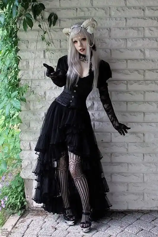 A person with long silver hair and small horns stands against a white brick wall, wearing a black gothic dress with lace…