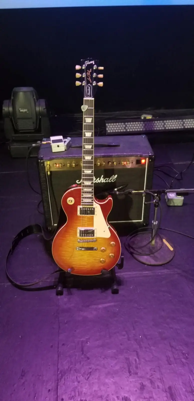 A sunburst Gibson Les Paul electric guitar stands upright on a stand on a purple stage floor, positioned in front of a black…