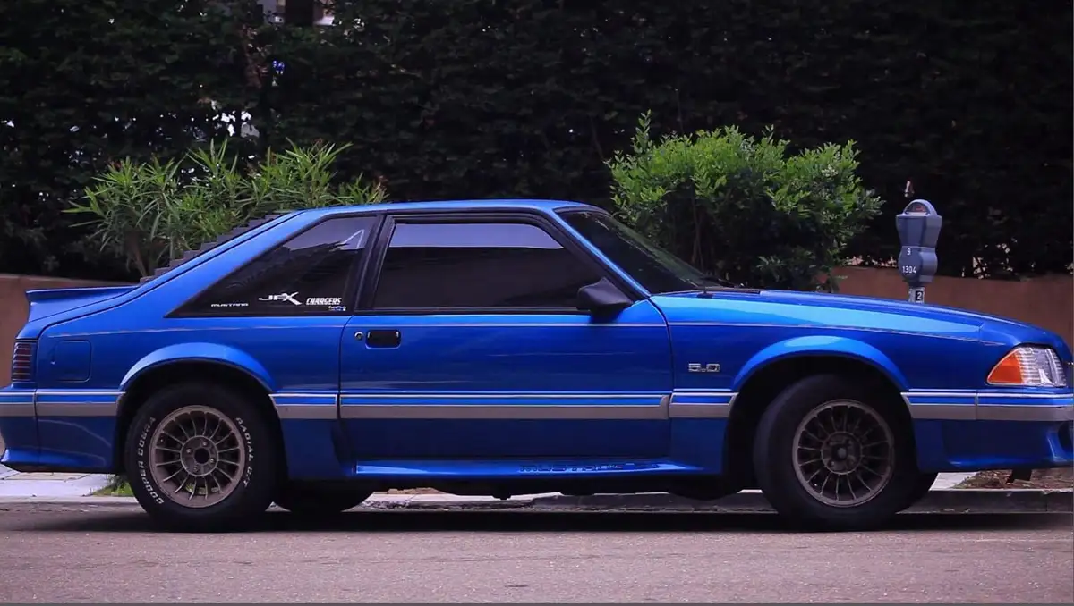 A bright blue 1980s Ford Mustang with silver racing stripes and aftermarket wheels is parked on a street next to a parking…