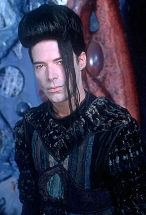 A man with a tall, black, pompadour-style haircut and a long, dark side-swept fringe stares intensely at the camera. He…