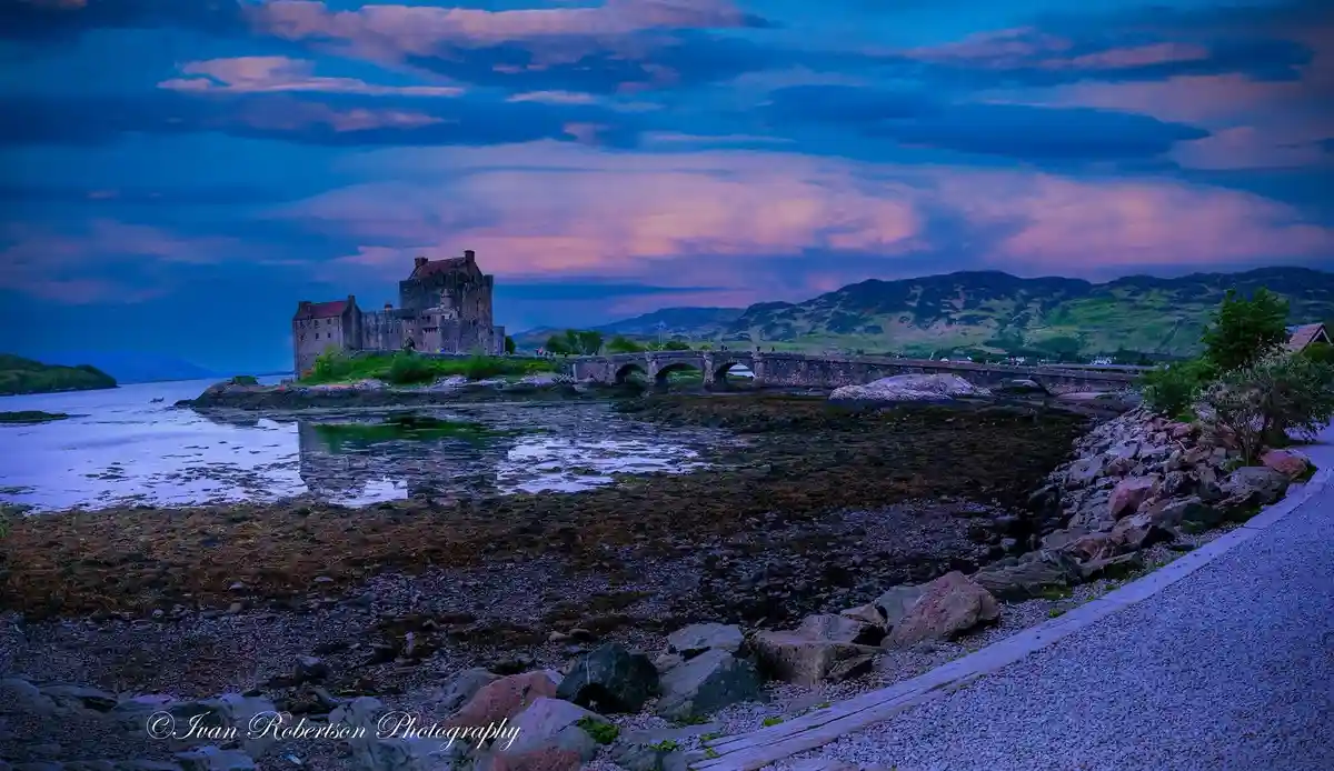 A scenic photograph of Eilean Donan Castle in Scotland at twilight, featuring a stone bridge connecting to the castle on an…