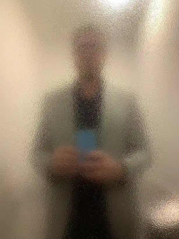 A blurry, distorted reflection of a person holding a phone is visible through a steamy, textured glass surface, likely a…