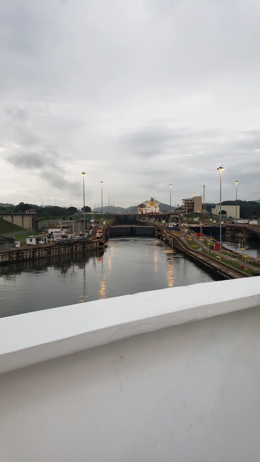 A wide-angle photo shows a large ship navigating through a canal lock system, likely the Panama Canal, under an overcast…