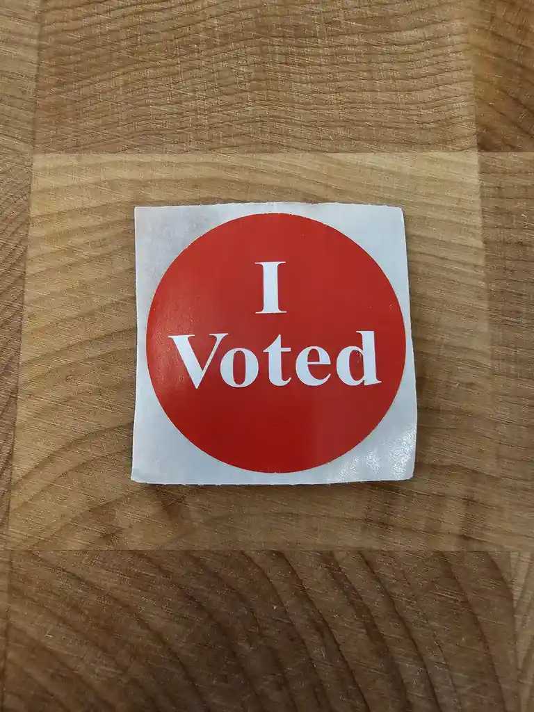 A red circular sticker with white text reading 'I Voted' is centered on a light brown wooden surface with visible grain…