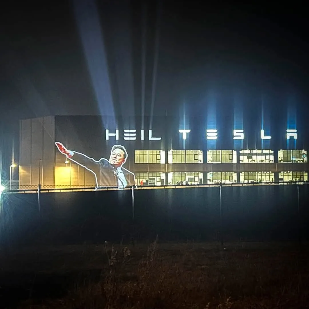 A nighttime photo of a large building with illuminated windows and spotlights. Projected onto the side of the building is a…