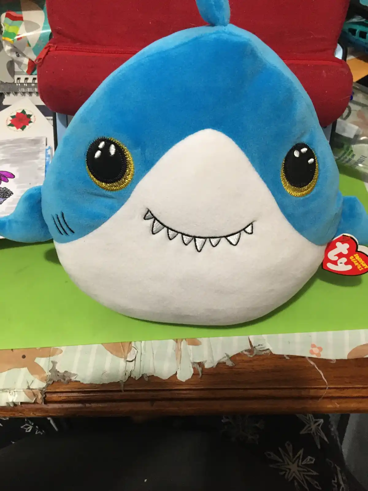A blue and white plush shark toy with large black eyes outlined in gold, a smiling mouth with small teeth, and a red Ty tag…
