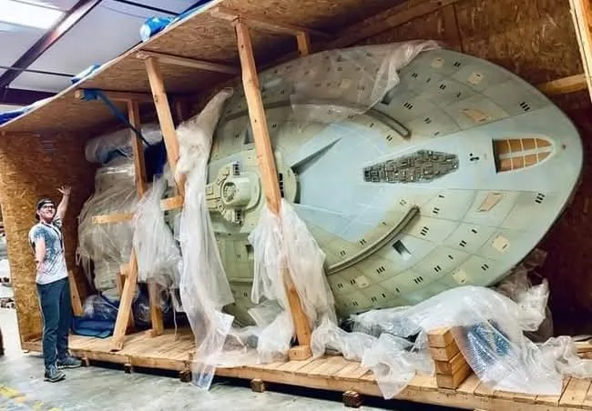 A person stands next to a massive, light-blue spaceship model, which is partially wrapped in plastic and secured with wooden…