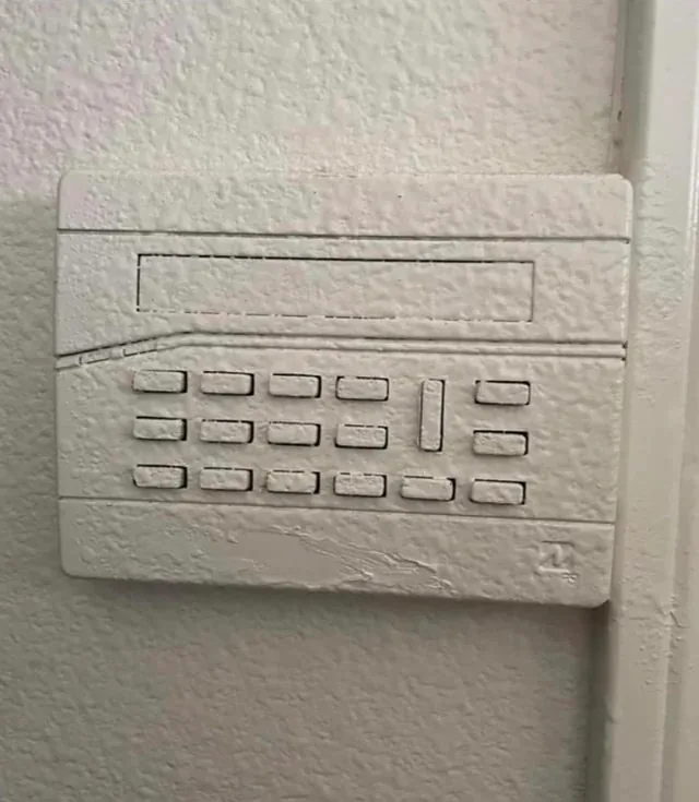 a close-up photo shows a security system keypad mounted on a wall, but the entire device has been painted over with white…