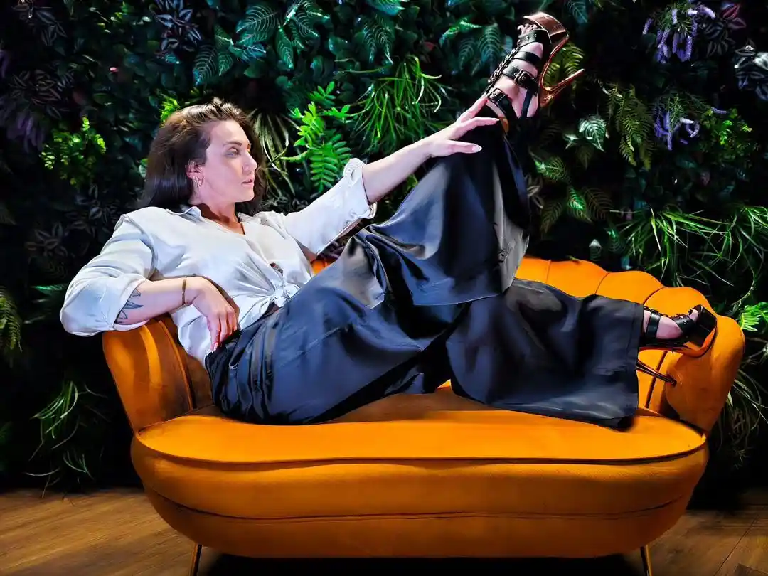 a woman lounges on a bright orange couch against a lush green foliage wall, wearing a white blouse and black wide-leg pants…