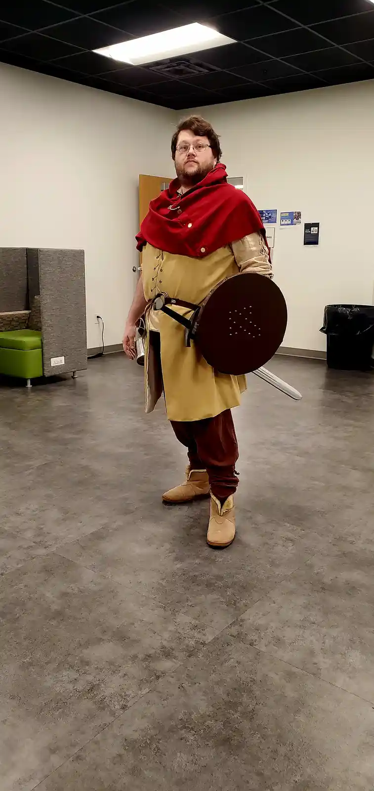 A man with glasses and a beard stands in a modern office space wearing a medieval-style costume with a red cloak, tan tunic…
