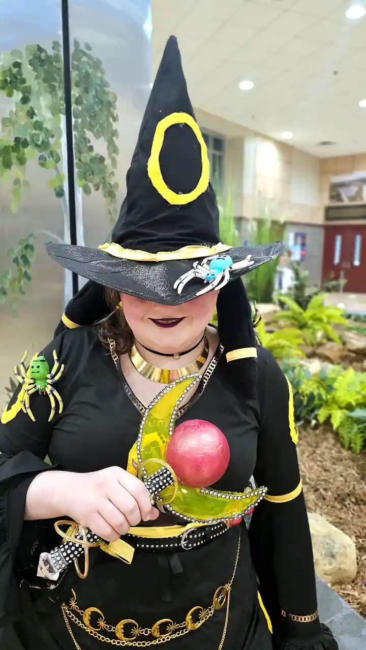 A person in a black witch costume with a tall pointed hat adorned with a yellow circle and a blue spider, smiling while…