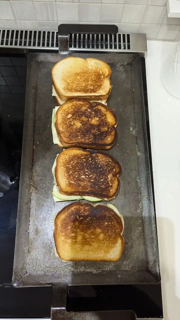Four sandwiches are cooking on a griddle, arranged vertically. The top sandwich is lightly toasted, while the three below it…