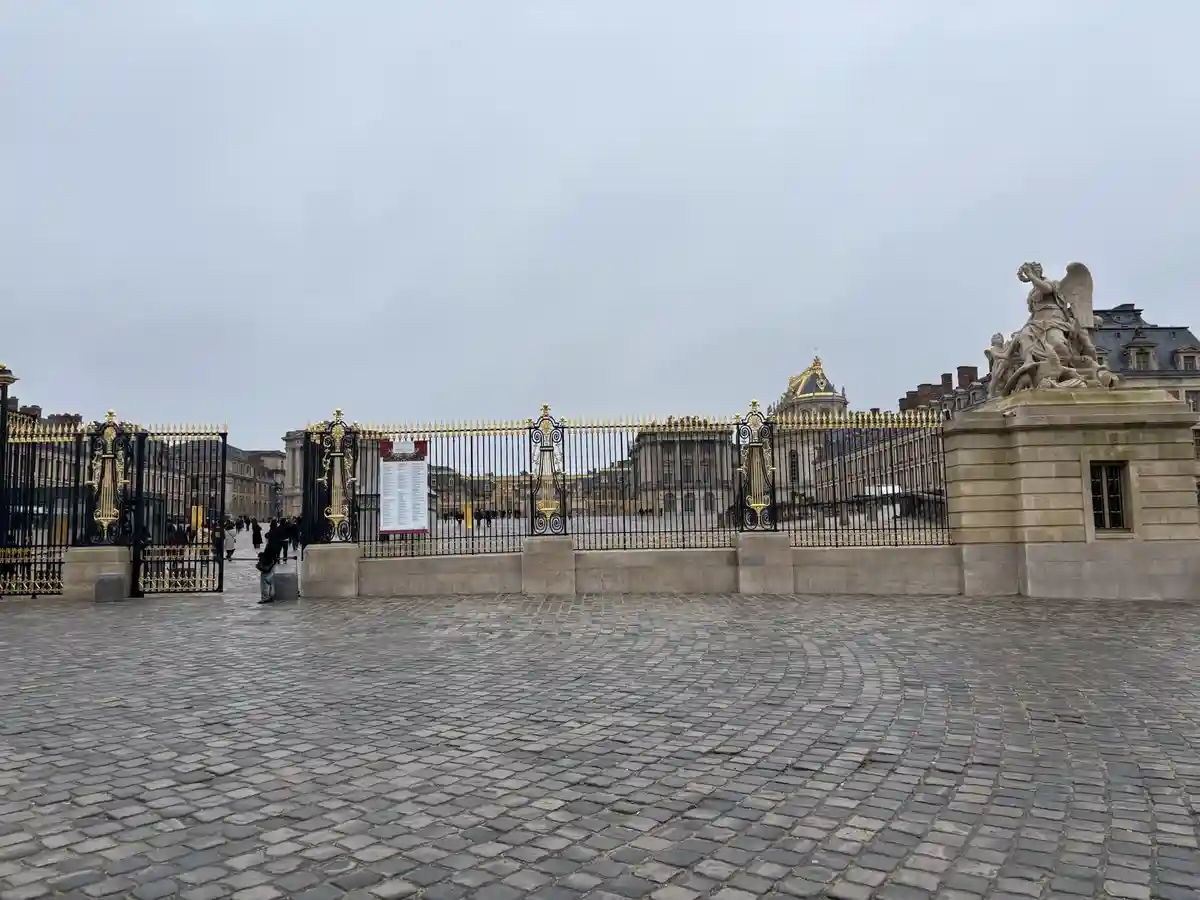 A wide-angle photo shows the ornate, black-and-gold gates of the Palace of Versailles under an overcast sky, with a large…