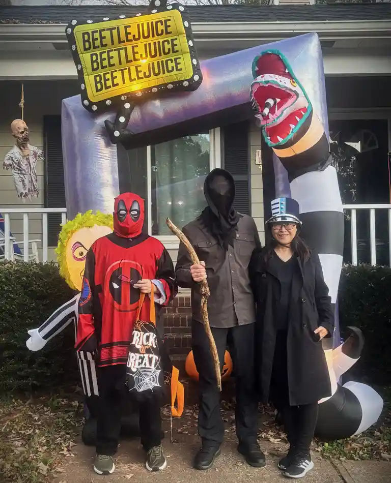 Three people pose in front of a large inflatable Beetlejuice-themed Halloween decoration. The person on the left wears a…