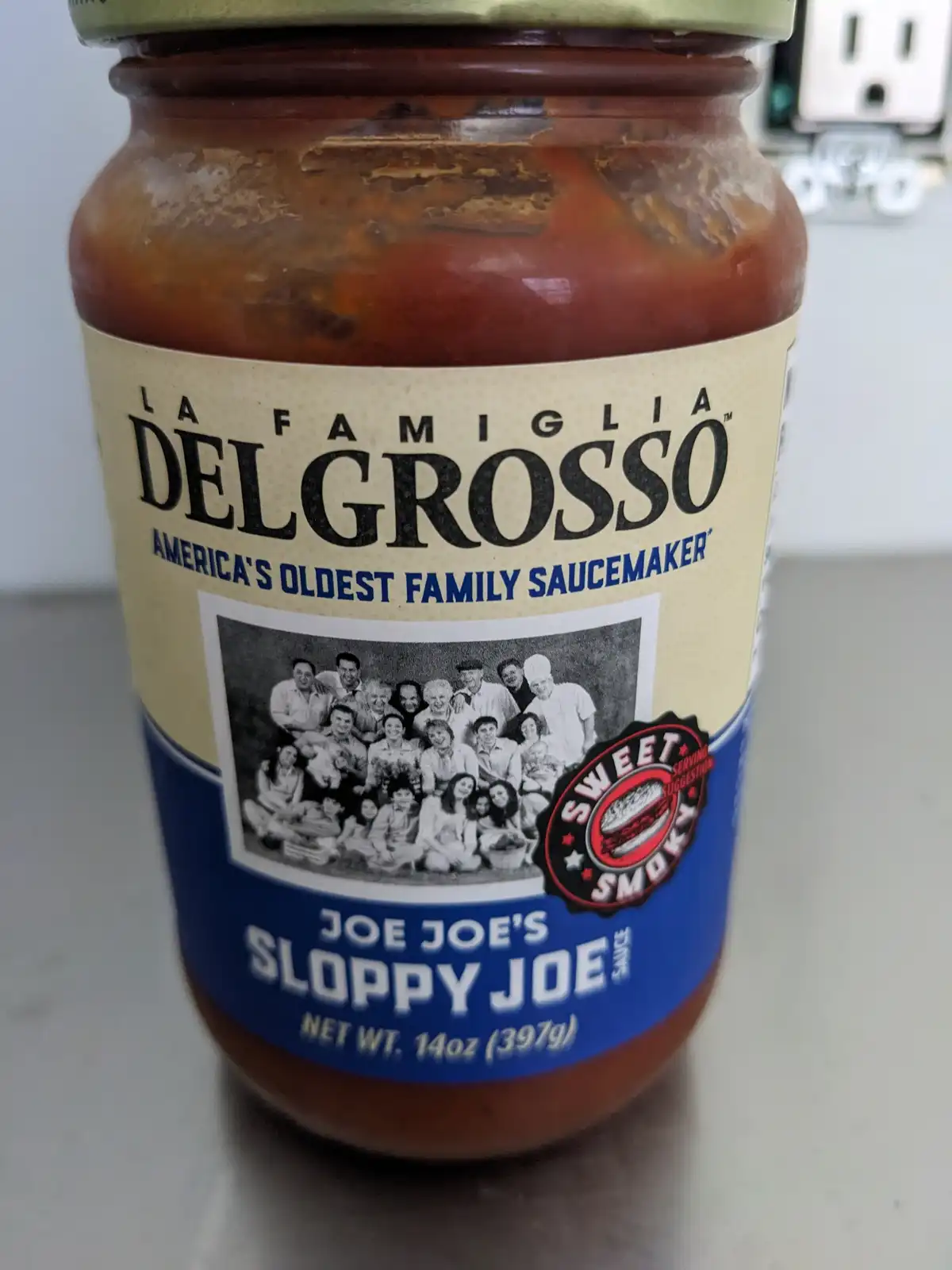 A close-up of a jar of La Famiglia DelGrosso 'Joe Joe's Sloppy Joe' sauce, featuring a family photo on the label and a…